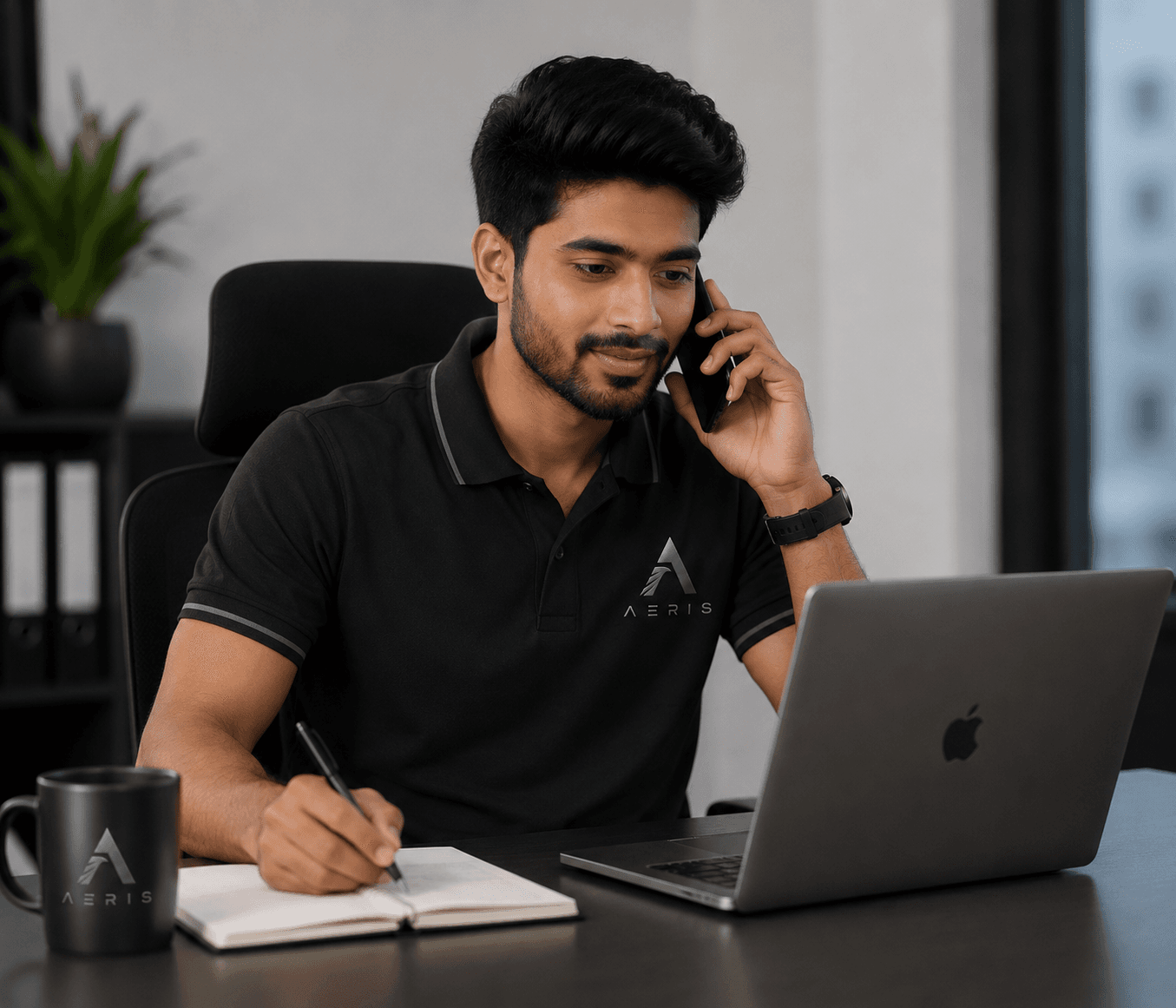 Man in black polo shirt talking on phone while writing notes and looking at laptop.