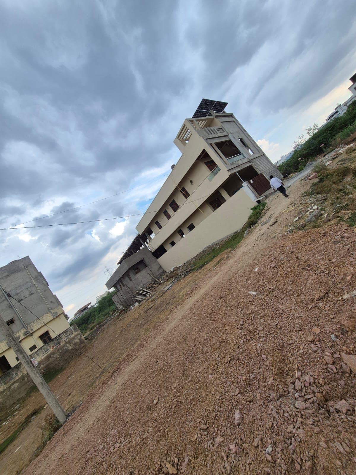 A modern building with solar panels sits beside a dirt road under a cloudy sky.