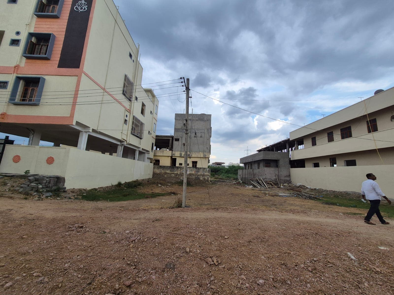 Man walking past multi-story buildings and a vacant dirt lot under a cloudy sky.