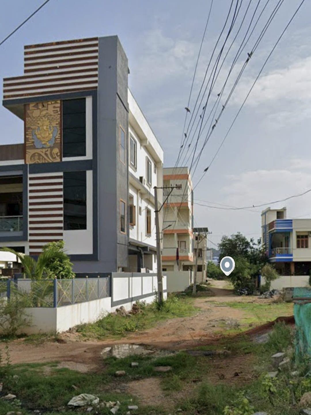 Modern multi-story building featuring a Ganesha mural on a dirt road with overhead power lines.